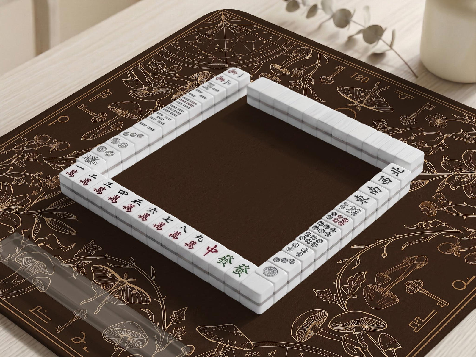 Dark brown mahjong table mat with a botanical border of moths, mushrooms, keys, vines, and small celestial details around a plain open center.