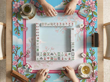 Floral cherry blossom mahjong mat in pink and sky blue with sakura branches and cloud details around an open center area.