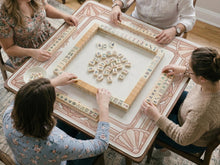Square blush pink mahjong mat with an art deco border, lotus motifs, and shell details on a light background.