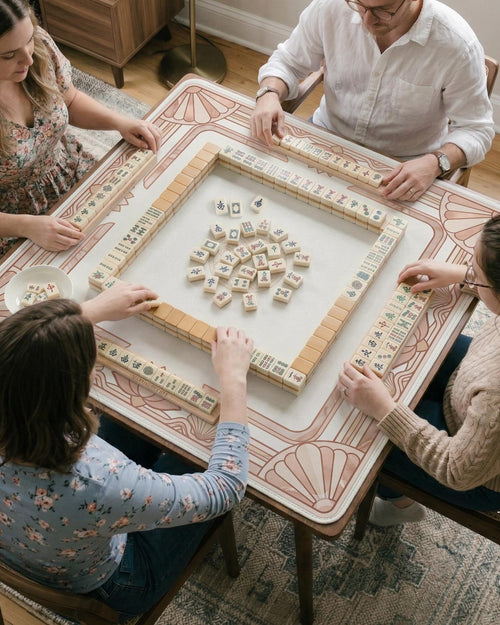 Square blush pink mahjong mat with an art deco border, lotus motifs, and shell details on a light background.