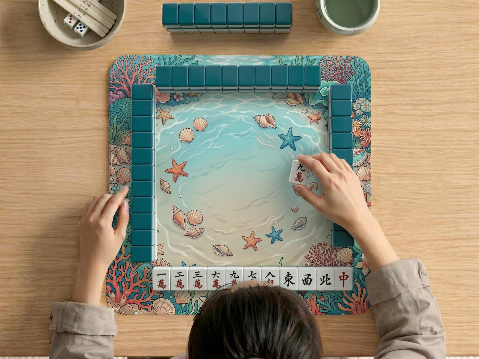 Square mahjong mat with coastal seashell artwork featuring coral, starfish, scallops, and aqua water colors around a lighter center area.
