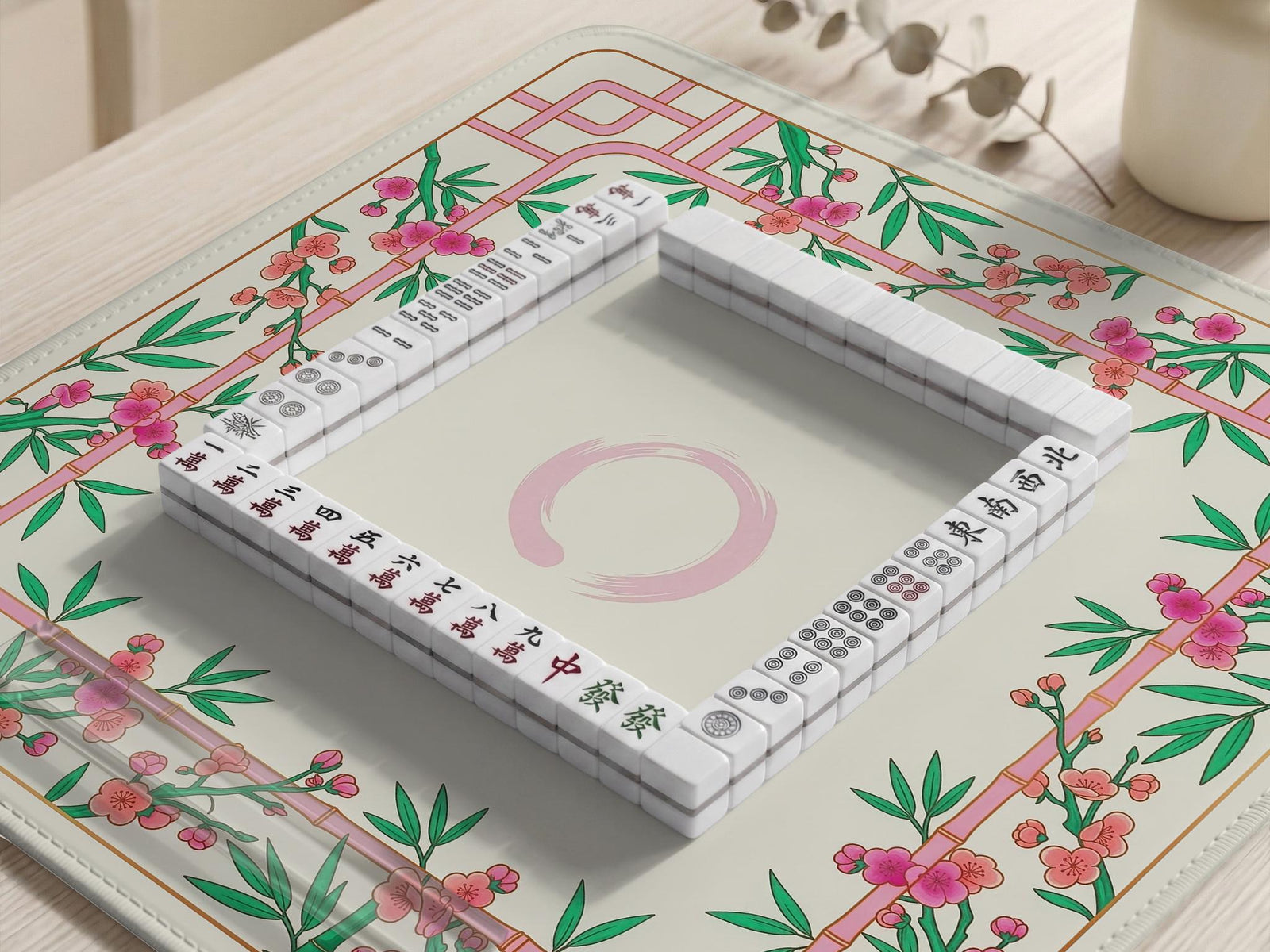 A square mahjong mat with a cream center, pink trellis pattern, green bamboo leaves, and small cherry blossom accents around the edge.