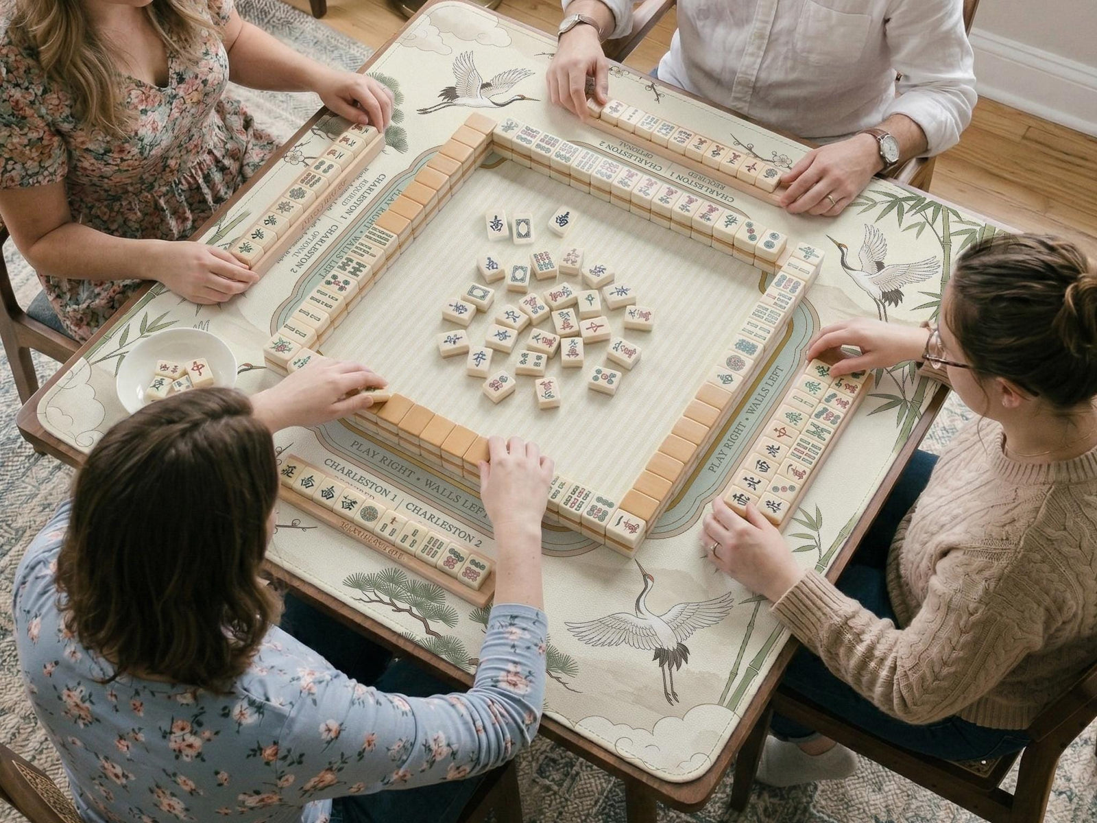 Sage green square mahjong mat with a chinoiserie design featuring cranes, bamboo, blossoms, and mountains, with rules printed around the border.
