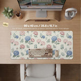 Floral desk mat with sage greenery, pink peonies, blue flowers, and a warm cream background.