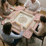 Square pink mahjong mat with a gingham border, floral corner details, and ribbon accents on a light background.