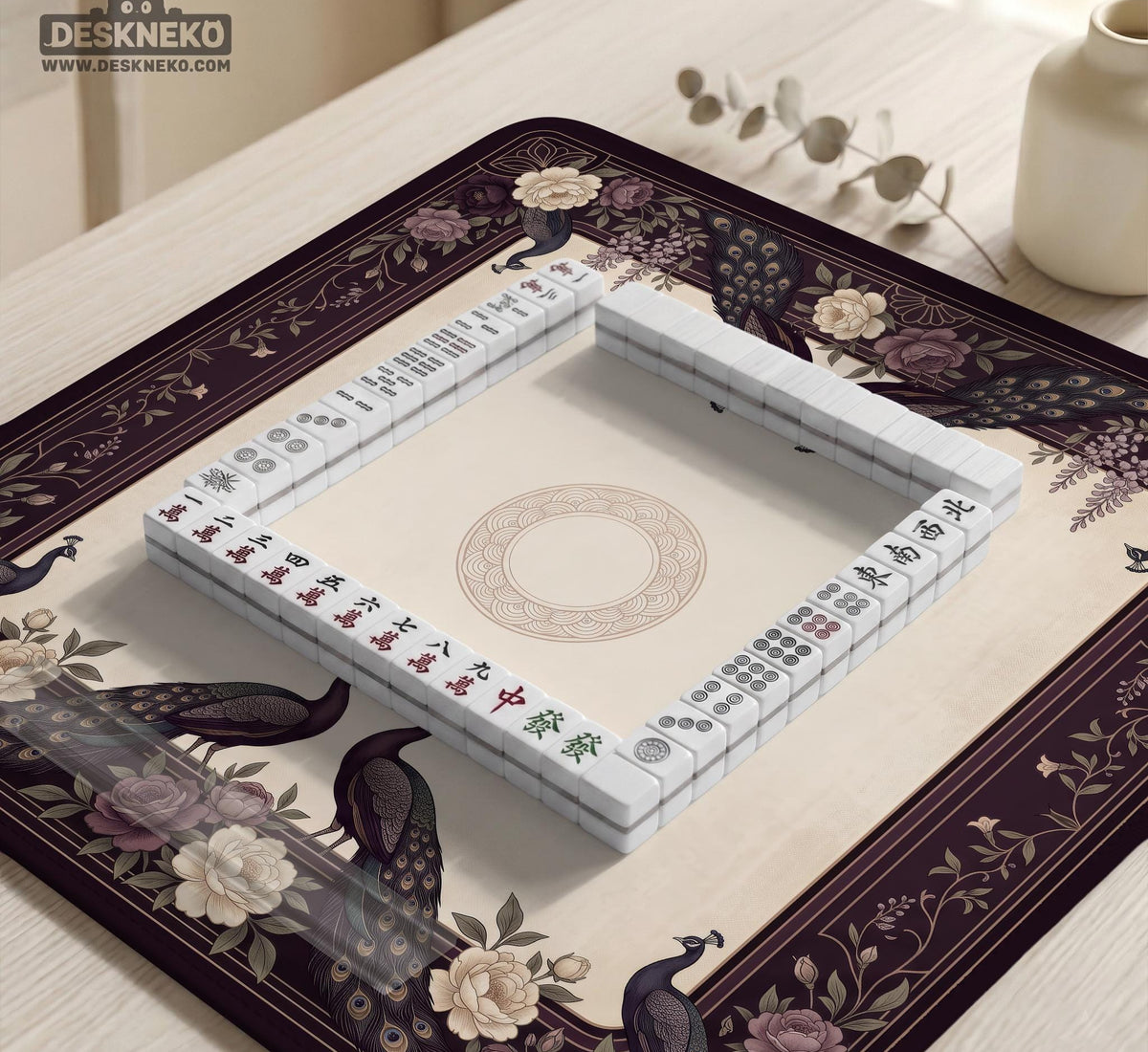 Square mahjong mat with a dark plum border, ivory floral pattern, muted sage leaves, and a decorative peacock garden design.