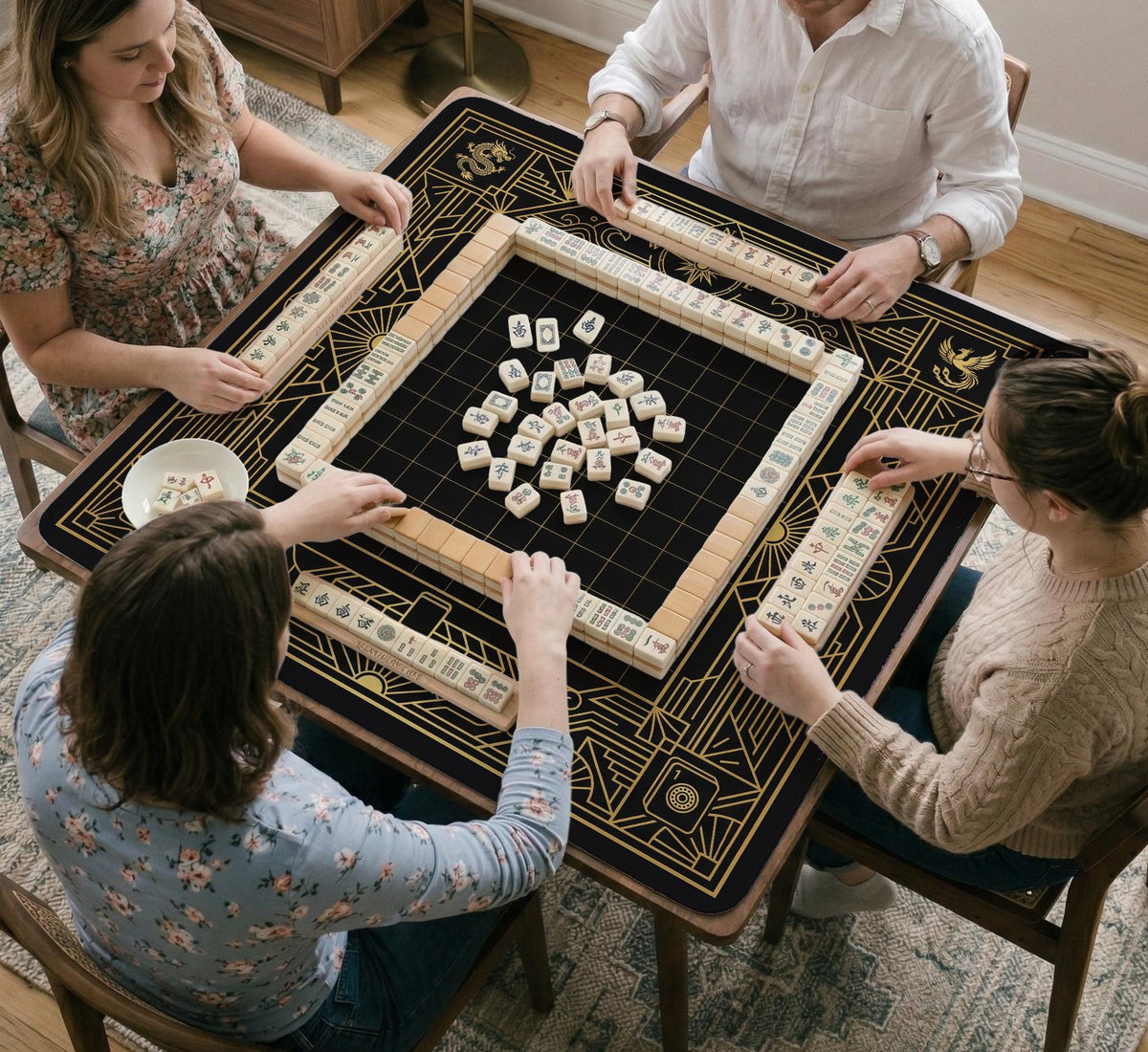 Black and gold square mahjong mat with a scalloped edge, geometric border, thin line details, fan motifs, and dragon and phoenix accents around an open center.