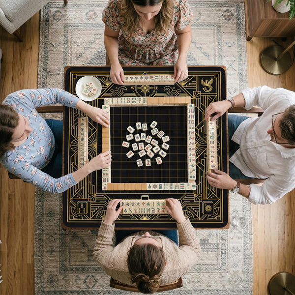 Black and gold square mahjong mat with a scalloped edge, geometric border, thin line details, fan motifs, and dragon and phoenix accents around an open center.