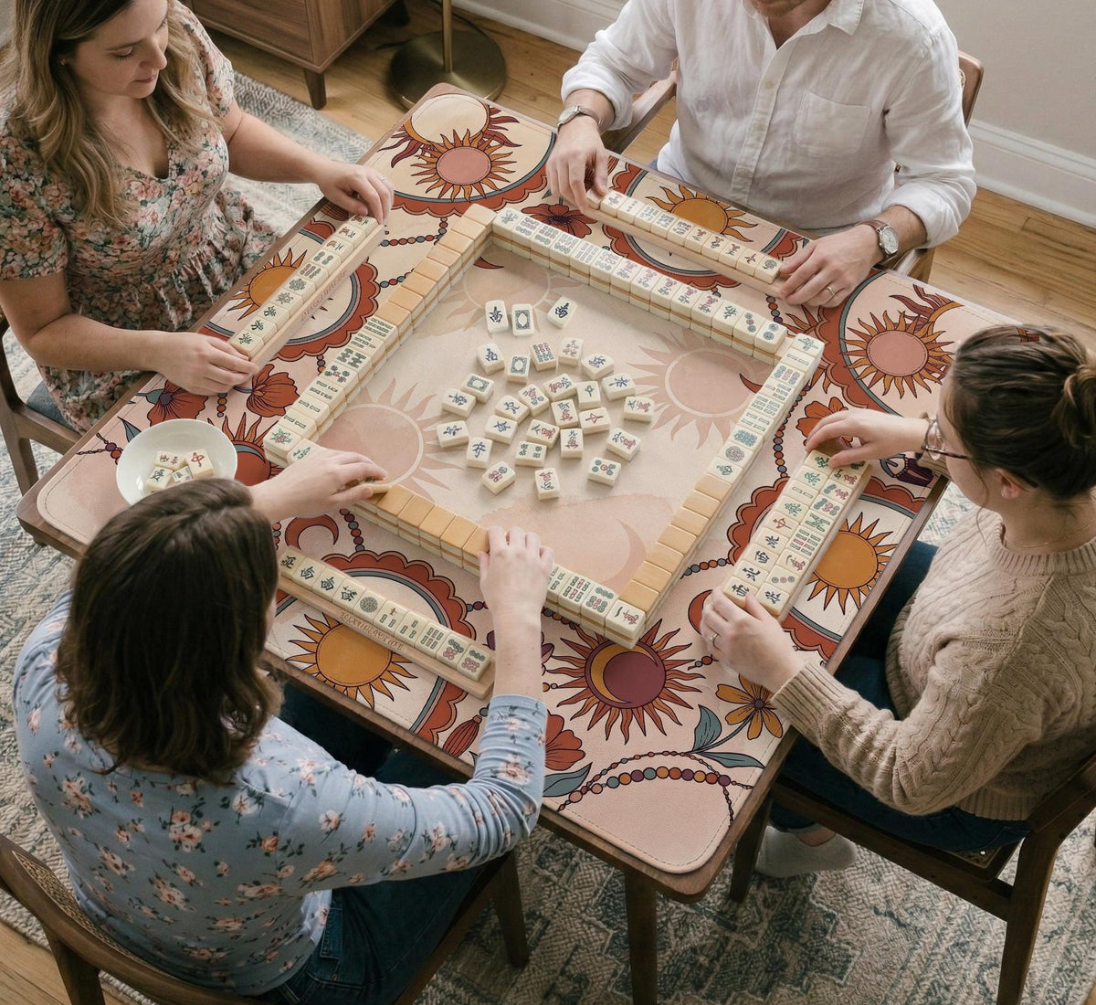 A square mahjong mat with a scalloped edge, sun and moon artwork, terracotta flowers, a soft cream background, and a decorative border around the play area.