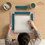 Pink gingham mahjong mat with a floral border, cherry blossom details, and a bow design around a plain center play area.