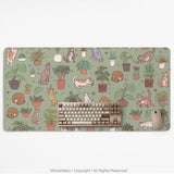 A desk mat with a repeating pattern of cats, leafy houseplants, cactus pots, and small plant-shop style details in sage green.