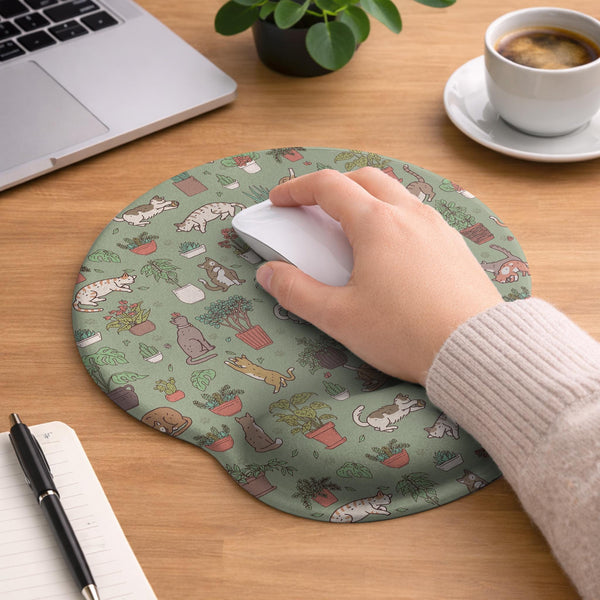 Sage green desk mat with a pattern of lounging cats, leafy plants, cactus pots, and earthy botanical details.