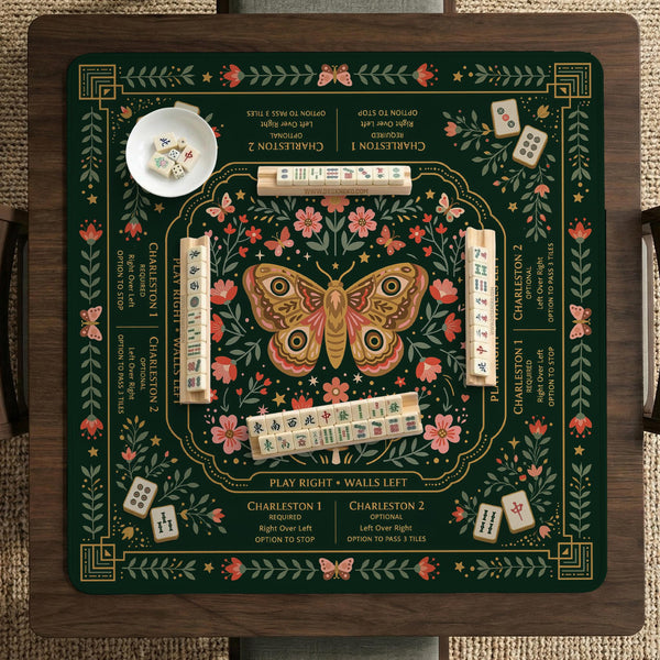 Dark botanical mahjong table mat with a floral border, large moth and butterfly design in the center, gold corner frame, and printed rules layout around the edge.