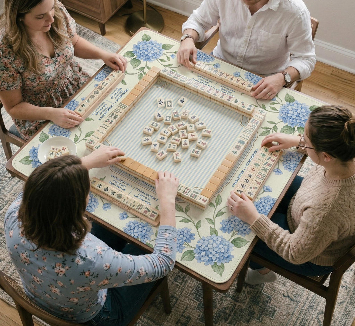 A square mahjong mat with blue hydrangea flowers, green vines, a cream background, a striped center, and printed rules around the border.