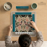 Beige mahjong mat with a botanical floral bird design, blush flowers, olive green leaves, black flower accents, and a warm linen-style background. Scalloped edge option shown.