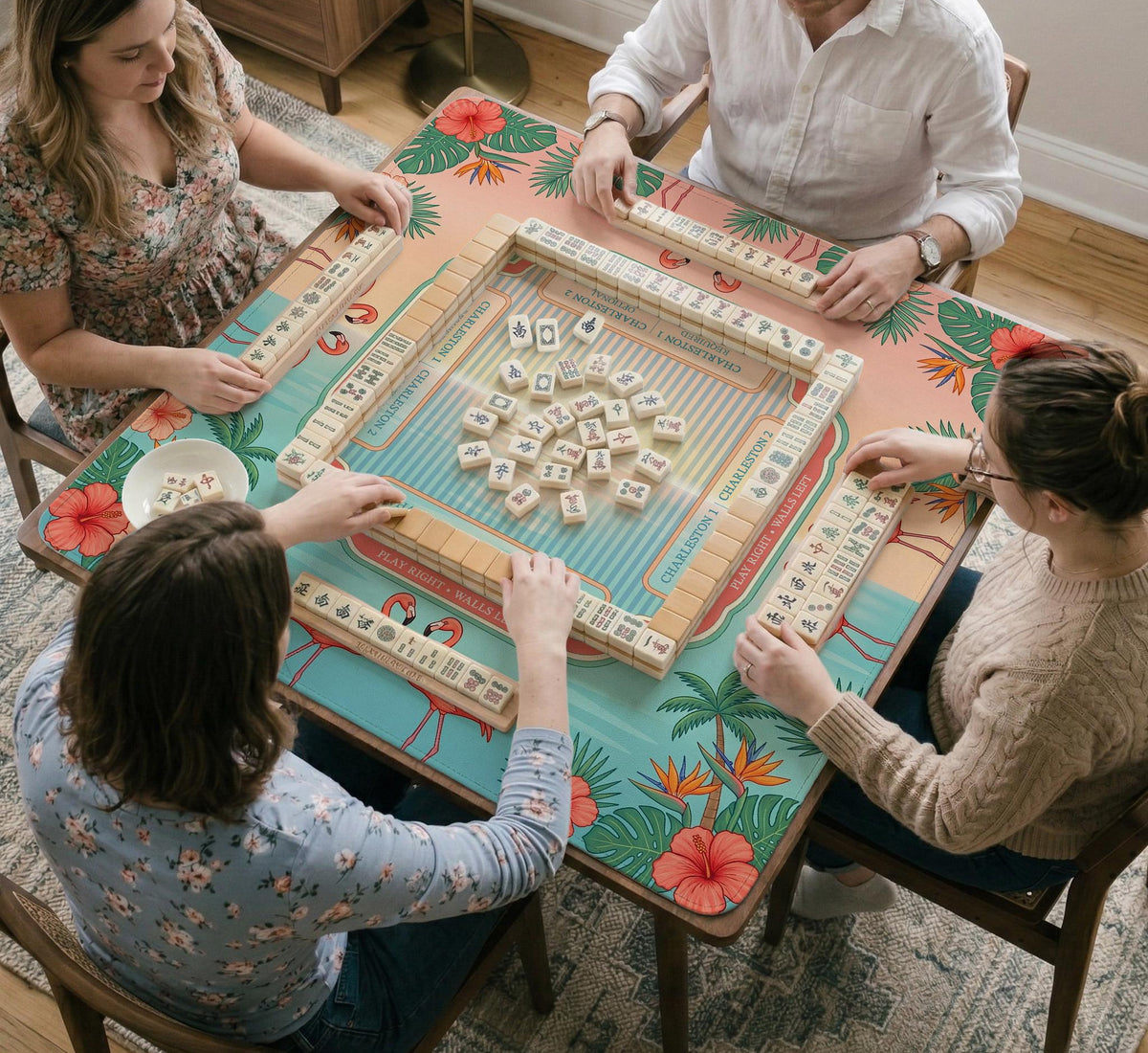 A square mahjong mat with flamingos, hibiscus flowers, palm leaves, and aqua water accents on a coral background with a scalloped edge.