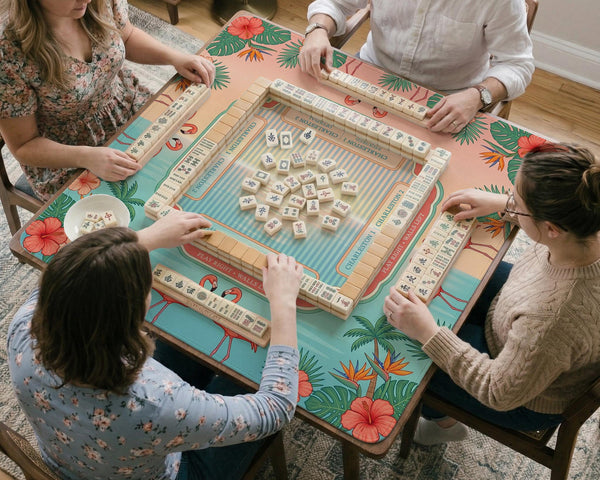 A square mahjong mat with flamingos, hibiscus flowers, palm leaves, and aqua water accents on a coral background with a scalloped edge.