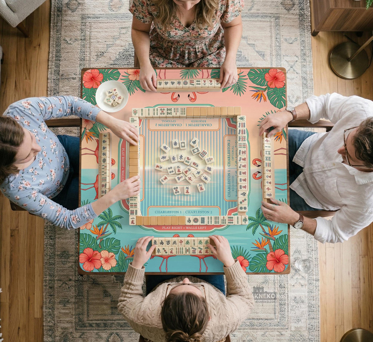 A square mahjong mat with flamingos, hibiscus flowers, palm leaves, and aqua water accents on a coral background with a scalloped edge.