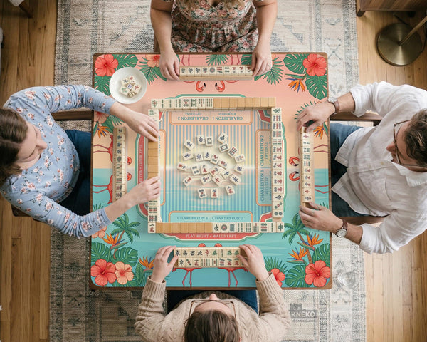 A square mahjong mat with flamingos, hibiscus flowers, palm leaves, and aqua water accents on a coral background with a scalloped edge.