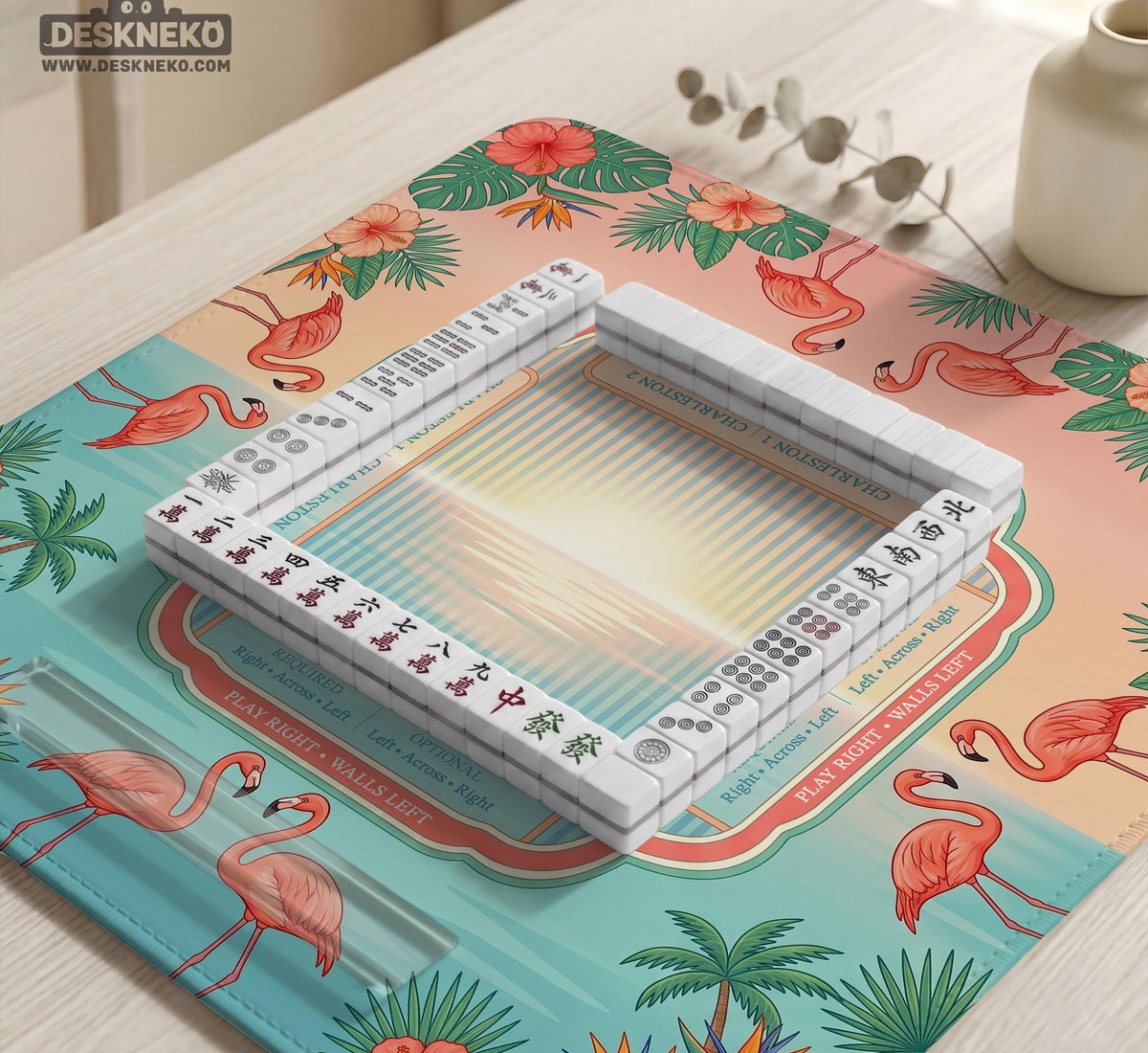 A square mahjong mat with flamingos, hibiscus flowers, palm leaves, and aqua water accents on a coral background with a scalloped edge.