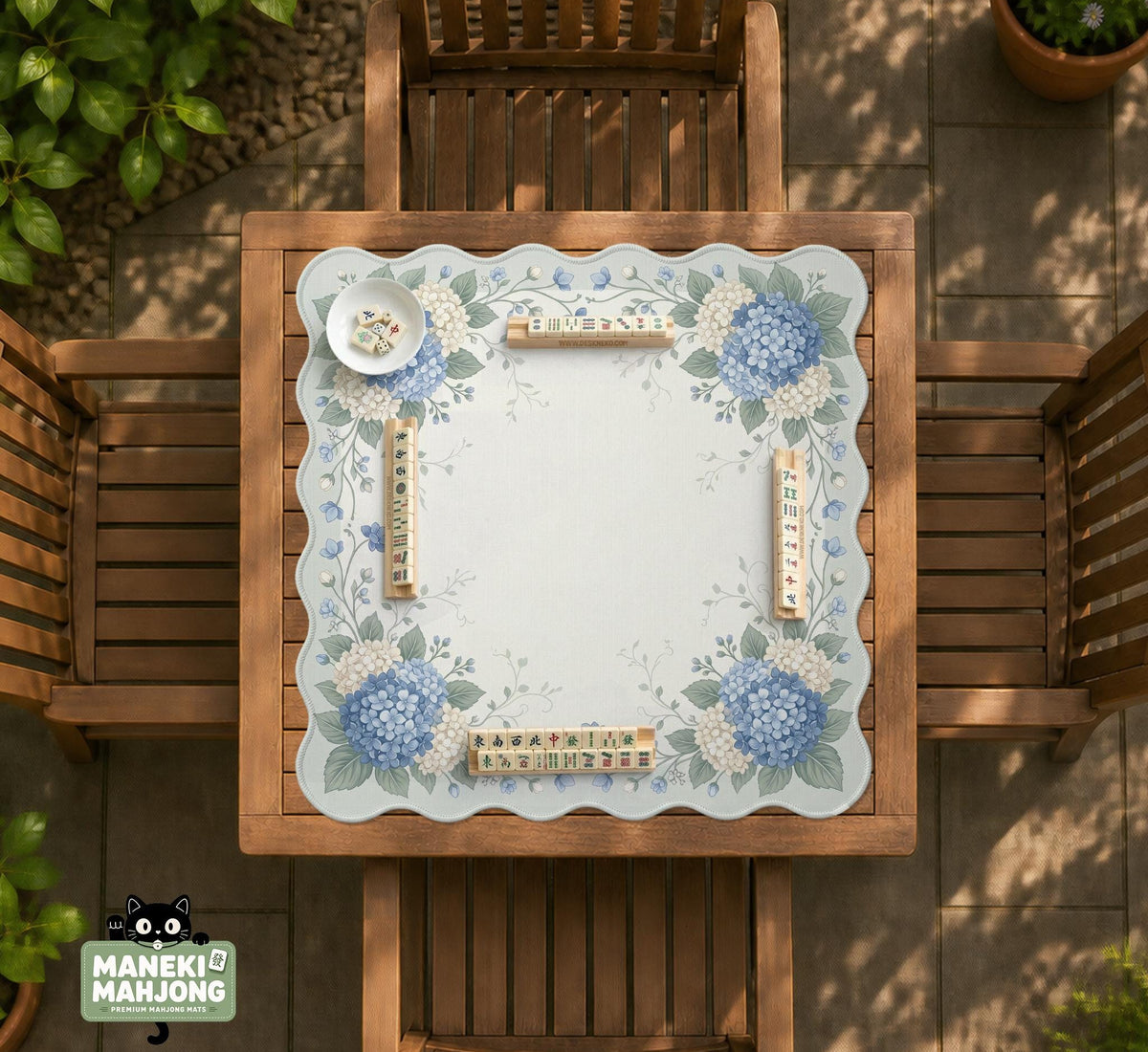 A square blue mahjong mat with hydrangea flowers, cream blossoms, sage leaves, and vines around a plain center play area. The edge is scalloped.