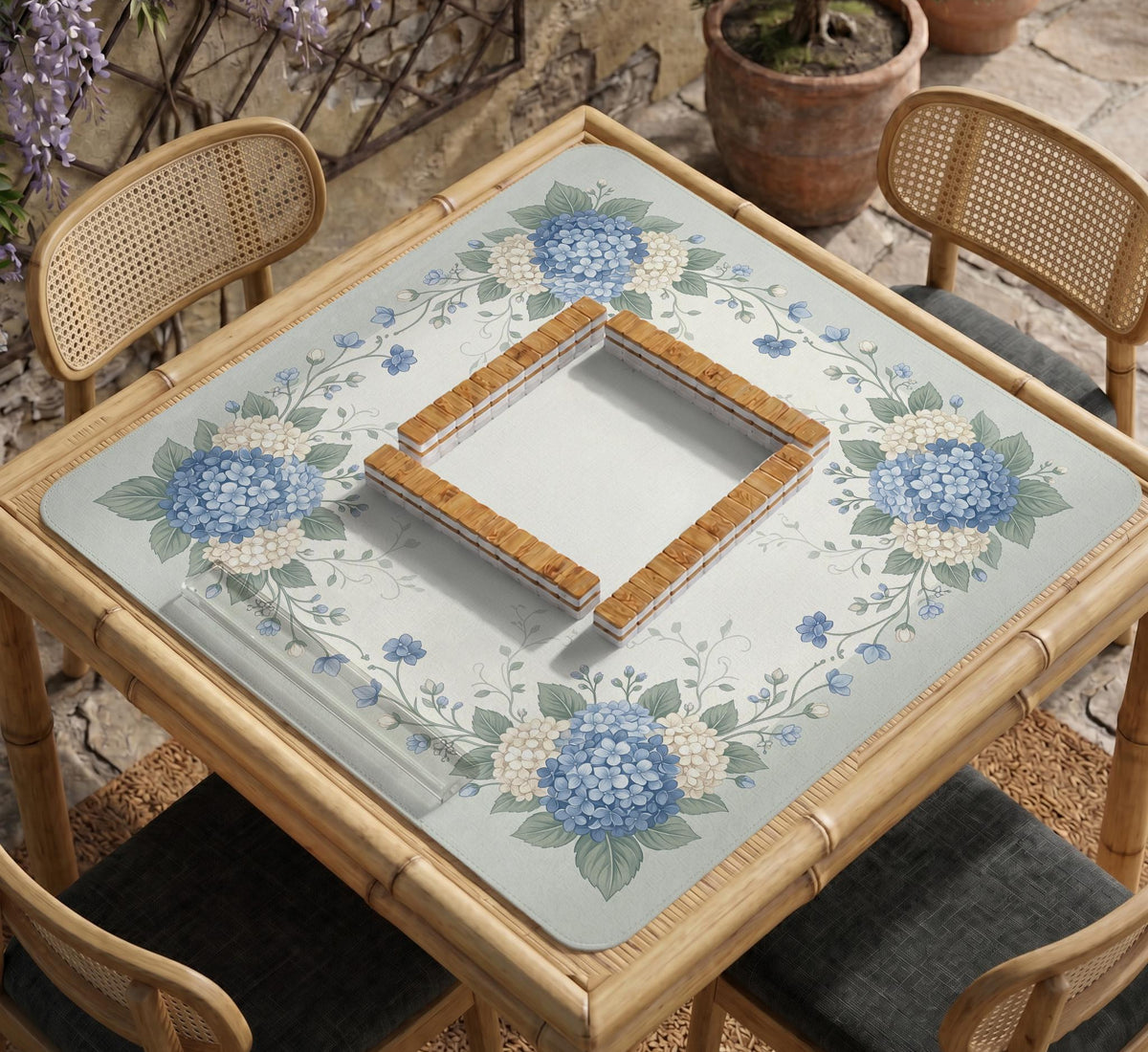 A square blue mahjong mat with hydrangea flowers, cream blossoms, sage leaves, and vines around a plain center play area. The edge is scalloped.