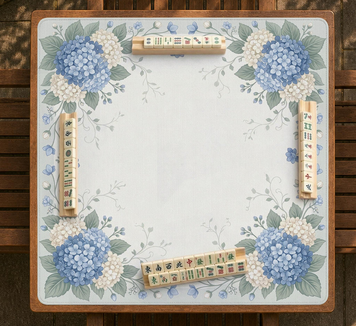 A square blue mahjong mat with hydrangea flowers, cream blossoms, sage leaves, and vines around a plain center play area. The edge is scalloped.